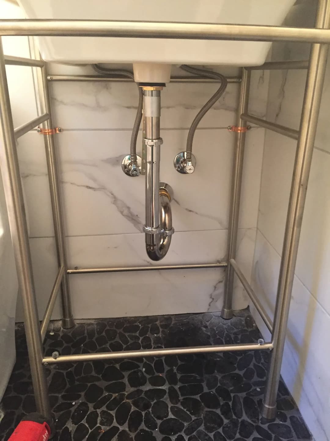 Under-sink plumbing with metallic pipes and a sink support frame on tiled floor.