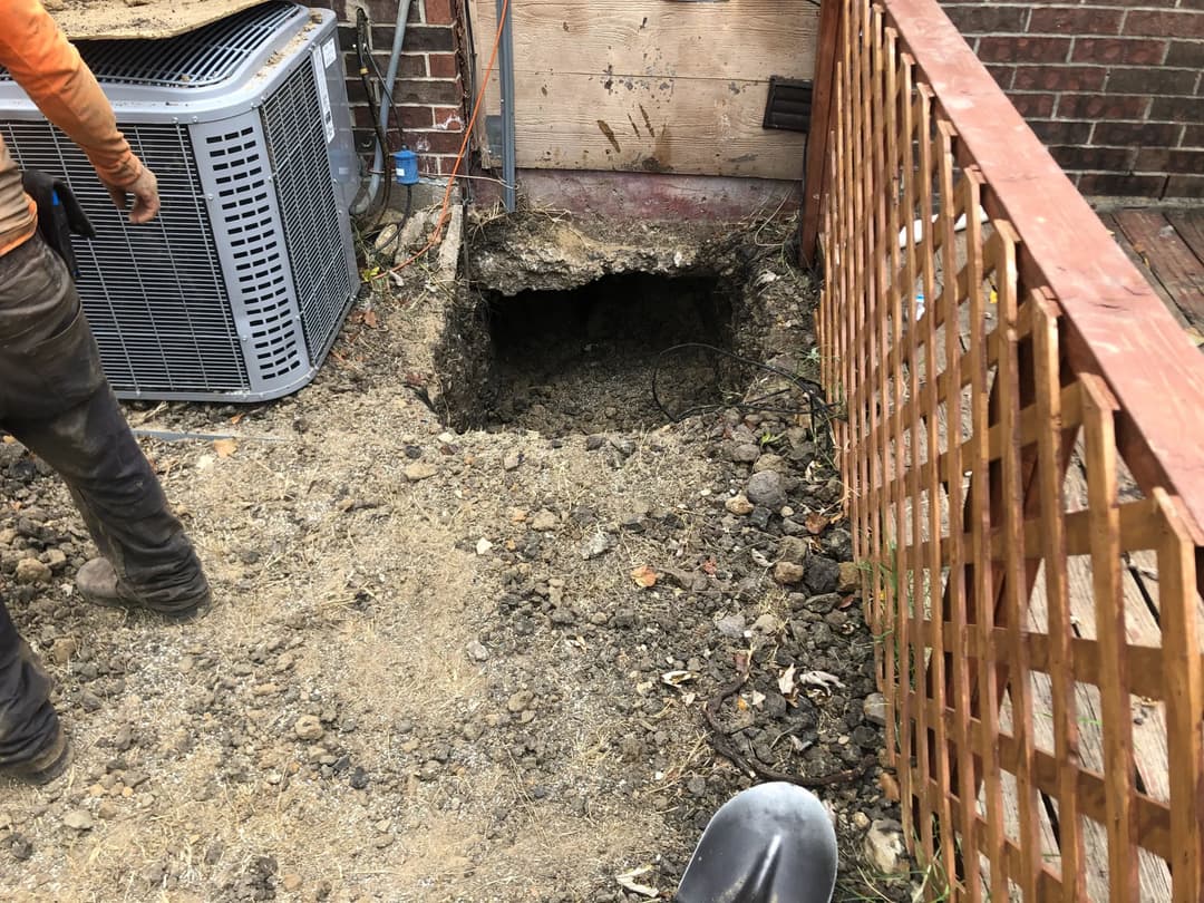 Excavated area beside air conditioning unit, showing dirt and tools for landscaping project.