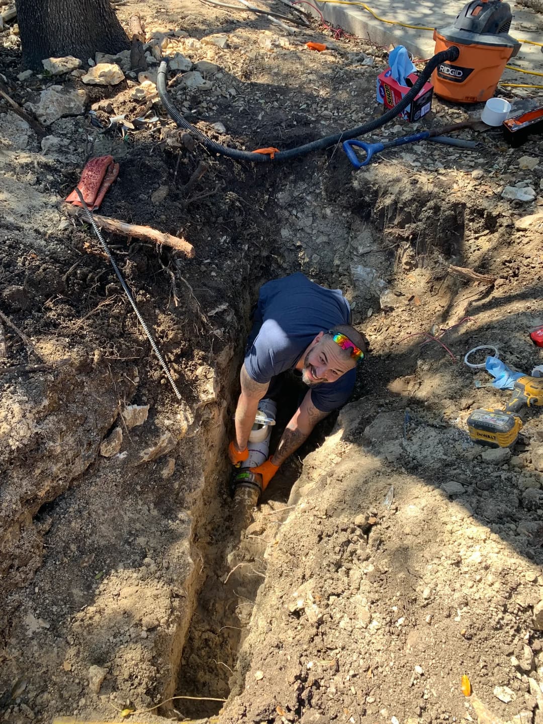 Person digging a trench in the ground with tools scattered around, outdoors on a sunny day.