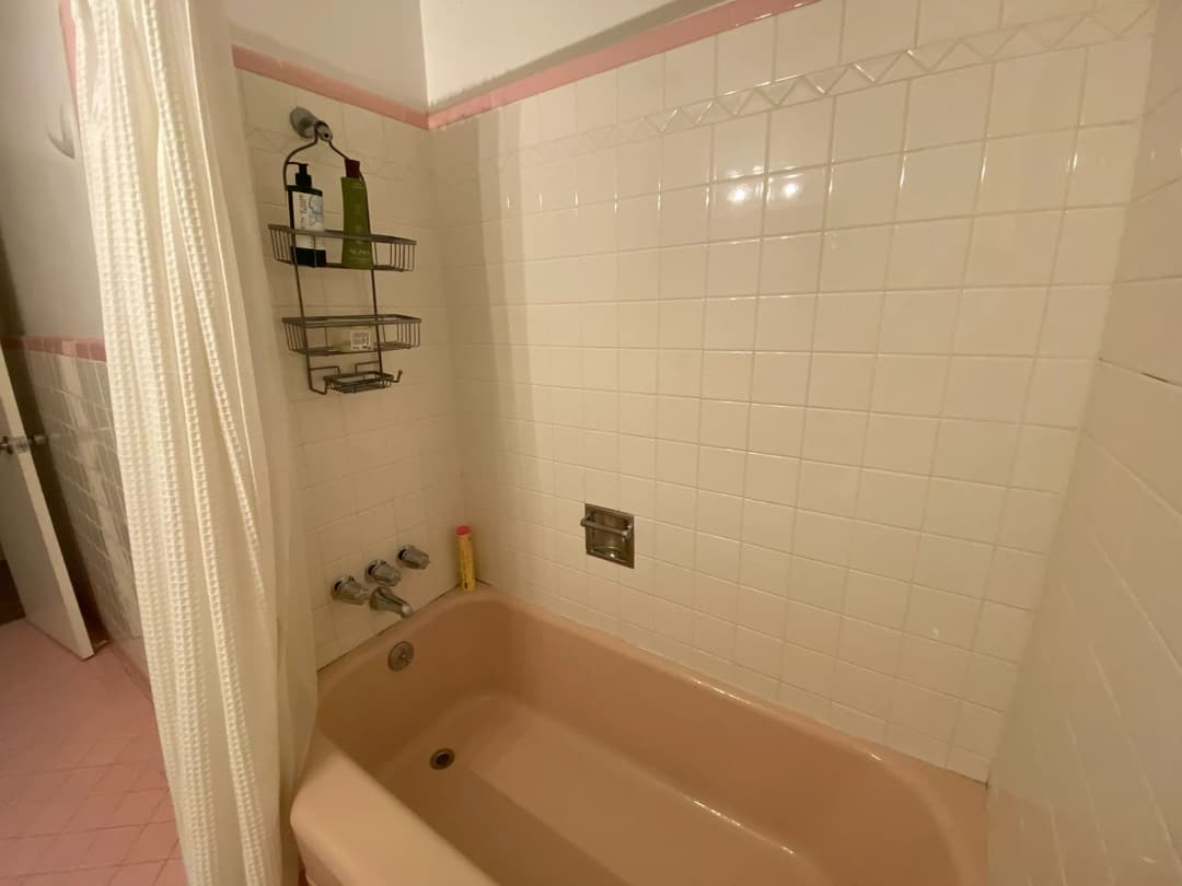 Pink bathtub in a tiled bathroom with shower fixtures and storage rack for toiletries.