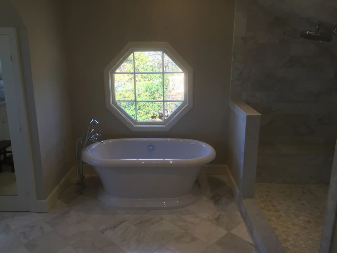 Elegant white bathtub in modern bathroom with octagonal window and stone shower.