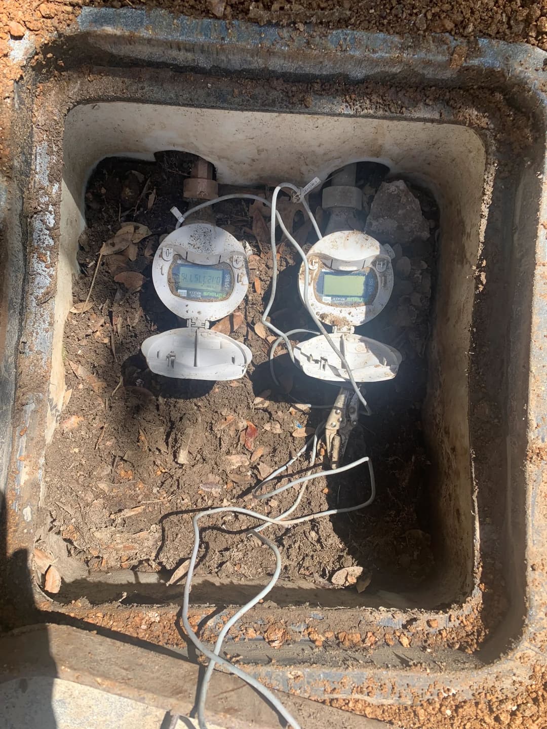 Two water meters in a dug-out pit with dirt and leaves around them.