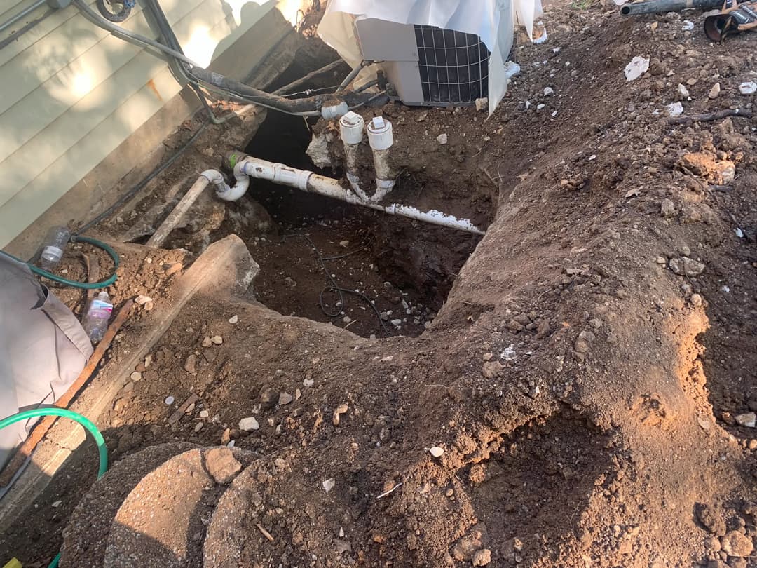 Excavated area next to building, revealing pipes and HVAC unit in a construction site.