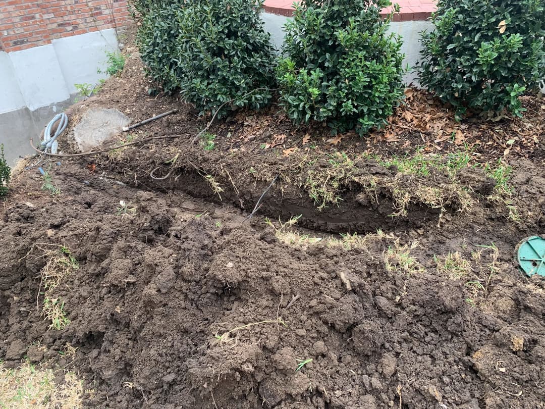 Trenched area in yard showing soil, grass, and shrubbery, with visible irrigation lines.